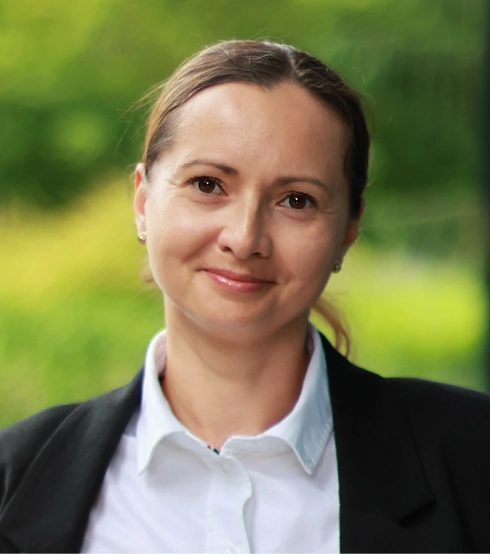 Natural Pharmaceuticals OmegaMarine Junior+™ Profesjonalny portret uśmiechniętej kobiety w białej koszuli i ciemnej marynarce na tle zieleni.