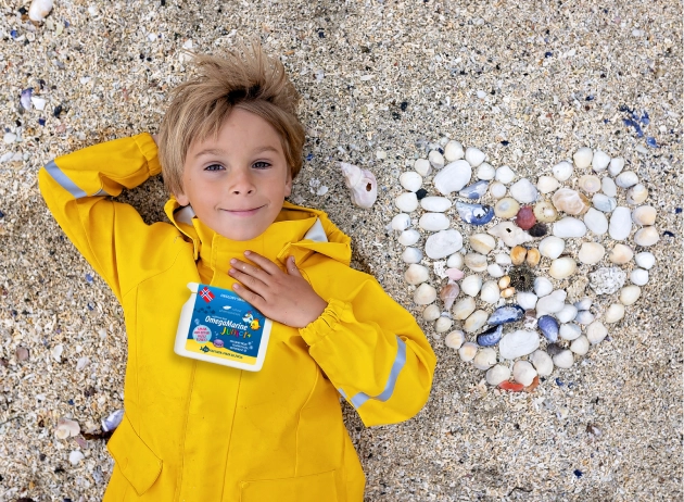 Natural Pharmaceuticals OmegaMarine Junior+™ Zdjęcie chłopca w żółtej kurtce, leżącego na plaży obok serca ułożonego z kamieni