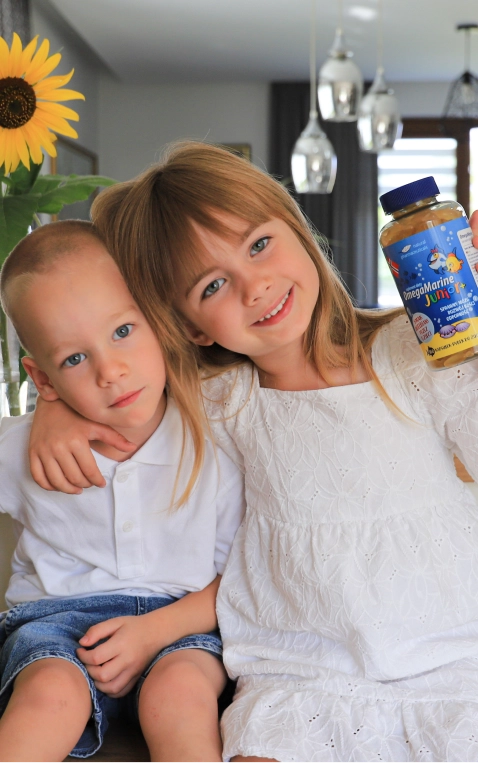 Natural Pharmaceuticals OmegaMarine Junior+™ Dwoje uśmiechniętych dzieci - dziewczynka trzymająca słoik z żelkami OmegaMarine Junior i chłopiec w jej objęciach, na tle słonecznika i jasnego wnętrza.