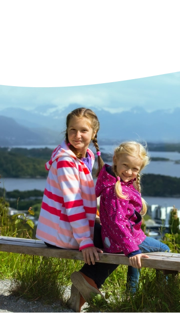 Natural Pharmaceuticals OmegaMarine Junior+™ Dwie uśmiechnięte dziewczynki siedzące na drewnianej ławce na wzgórzu z widokiem na jezioro i góry w tle. Starsza dziewczynka ma warkocze i nosi pasiastą bluzę w różowo-białe paski, młodsza ma blond włosy i różową kurtkę w kropki.