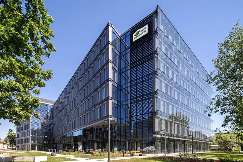 Natural Pharmaceuticals OmegaMarine Junior+™ Nowoczesny budynek biurowy Roche z przeszkloną fasadą w otoczeniu zieleni i błękitnego nieba.