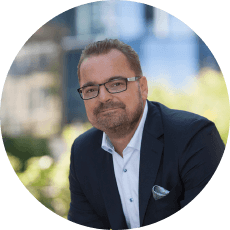 Natural Pharmaceuticals OmegaMarine Junior+™ Portret uśmiechniętego mężczyzny w okularach i marynarce na rozmytym miejskim tle.